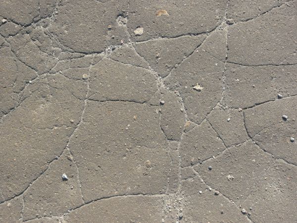 Asphalt texture with a rough, grey, cracked and uneven surface, and a few small white rocks on it.